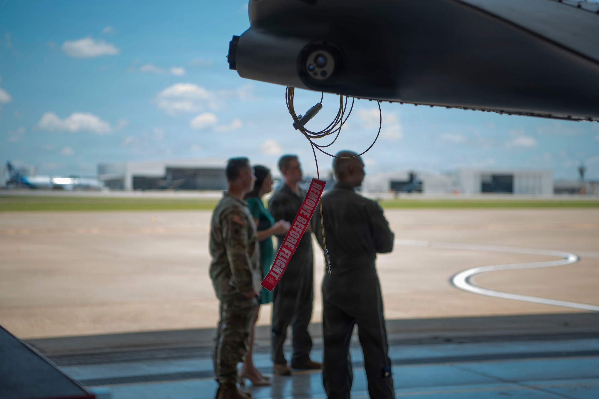 A remove before flight tag is attached to the wing of a plane as a group of people stand in the background in front of an airfield with green grass and blue sky