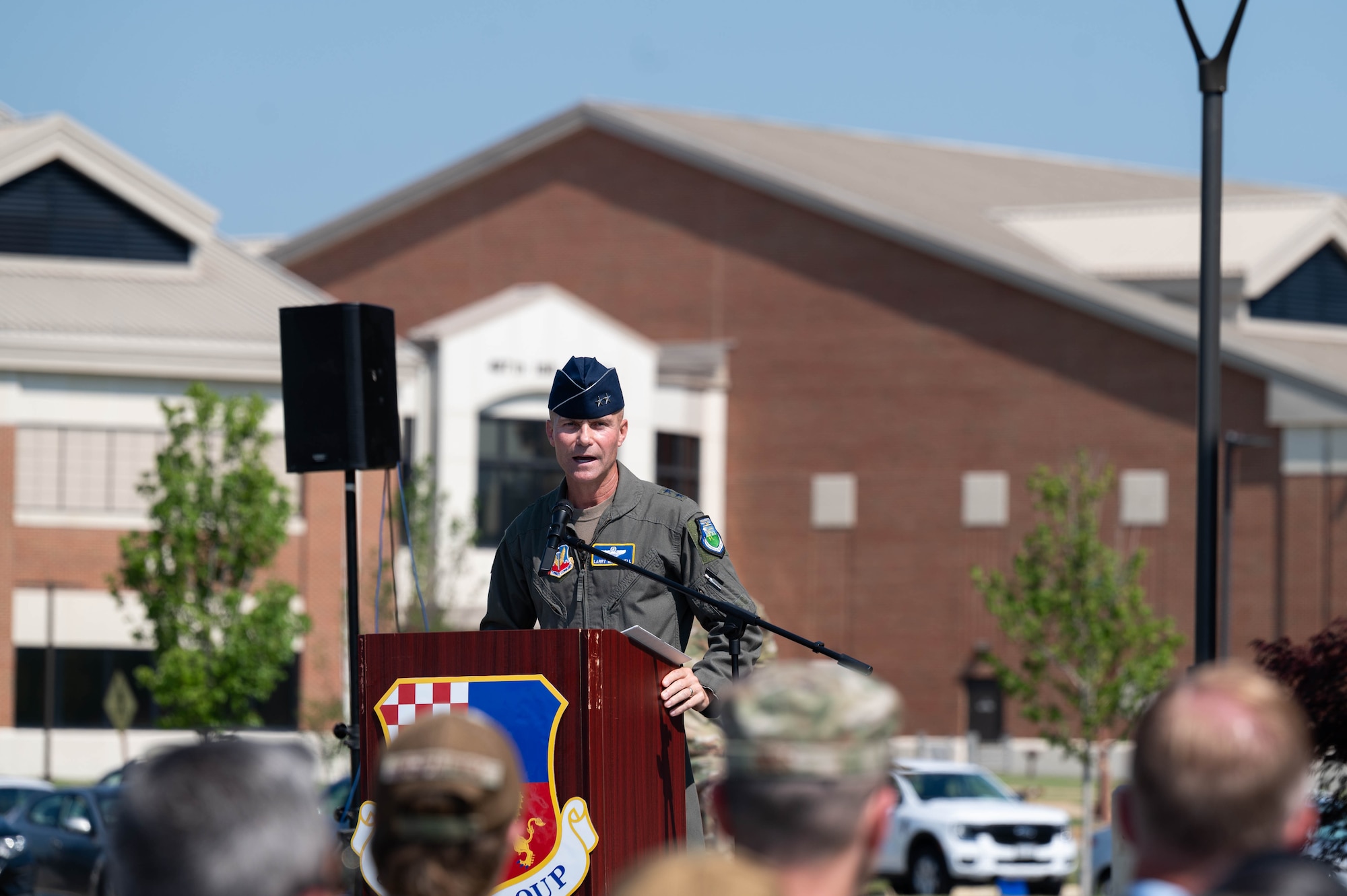 A U.S. Air Force officer stands at a podium and speaks to a crowd with buildings behind him.