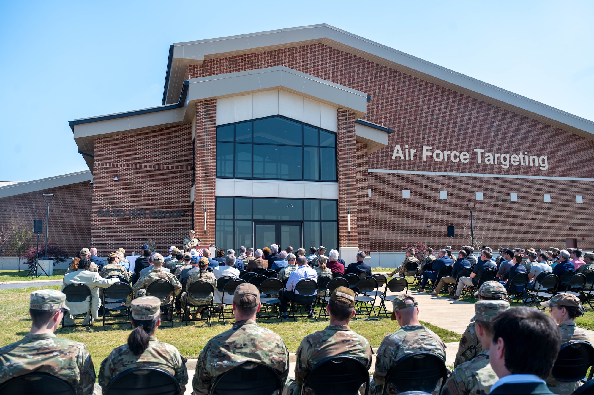 Military members and civilians sit outside in an audience in front of a newly constructed building. A military member is at a podium speaking.