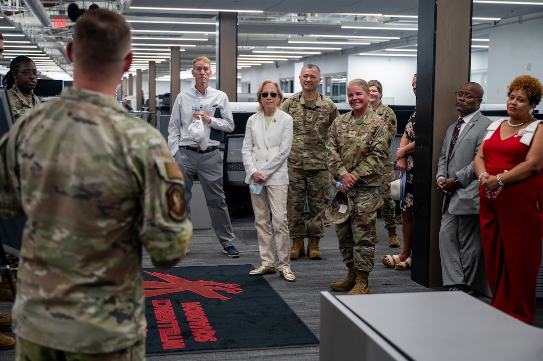 A group of Air Force members and civilians stand in a room, looking at an Airman briefing them.