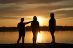 Three silhouettes standing in front of water and a sunset