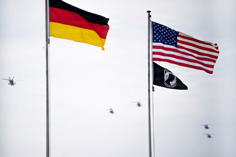 Left to right: the German flag, the American flag and the POW/MIA flag fly on flagpoles against a cloudy sky, as five helicopters fly in the distance.