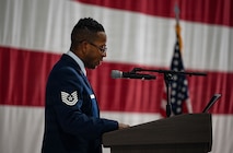 At a side angle, an Airman stands behind a podium giving his remarks. A U.S. flag is displayed behind him.